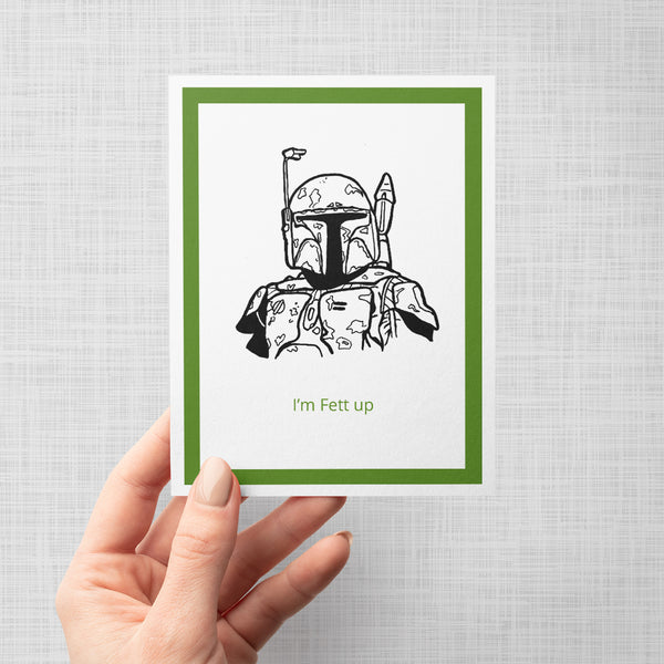 A hand holding a greeting card featuring a black line art illustration of Boba Fett from Star Wars, with the text 'I'm Fett up' printed in green below. The card is framed with a white border and a green inner border, set against a gray background.