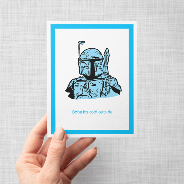 A hand holding a greeting card featuring a stylized blue and black illustration of Boba Fett with the text 'Boba it's cold outside' below, set against a white background with a bright blue border, shown on a light gray backdrop.