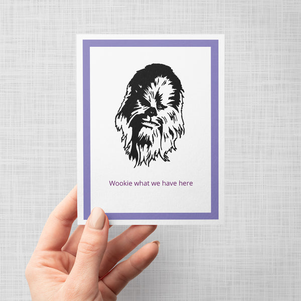 A hand holding a greeting card featuring a black line art illustration of Chewbacca on a white background, framed by a double purple border. Below the illustration, the text 'Wookie what we have here' is written in purple.
