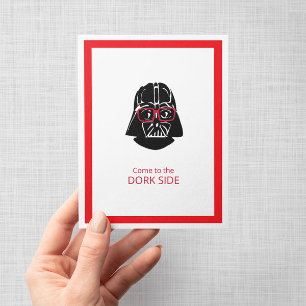 A hand holding a greeting card featuring an illustration of Darth Vader wearing red glasses, with the text 'Come to the DORK SIDE' below. The print has a white background, a red inner border shown against a neutral light gray background.