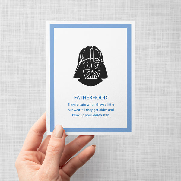 A hand holding a greeting card featuring a Darth Vader illustration above blue text 'FATHERHOOD. They're cute when they're little but wait 'til they get older and blow up your death star' set within a blue bordered frame on a light gray background. 