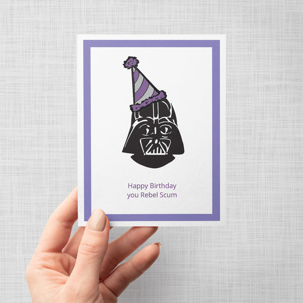 A hand holding a greeting card with a Darth Vader illustration wearing a party hat and text 'Happy Birthday you Rebel Scum' on a white background with purple border.