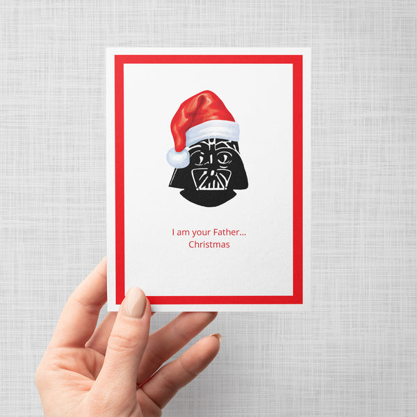 A hand holding a greeting card with a white center, a bright red border, and a stylized Darth Vader face wearing a red Santa hat. Below the illustration, red text reads, 'I am your Father Christmas on a gray background.