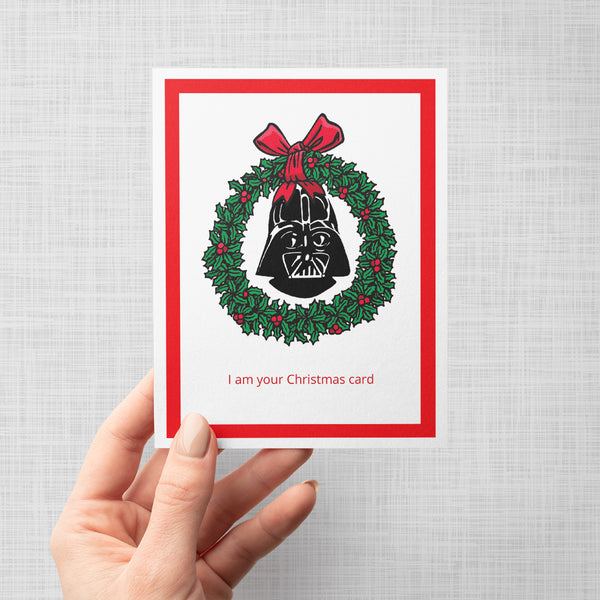 A hand holding a Christmas greeting card featuring Darth Vader's helmet inside a green holly wreath with a red bow, on a white card with a red border and the text 'I am your Christmas card' at the bottom.