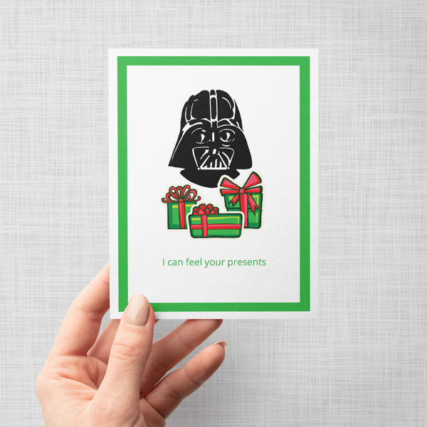 A hand holding a greeting card featuring a black Darth Vader head above three green gift boxes with red bows, with the text 'I can feel your presents' in green at the bottom. The card has a white center, a green border, and is shown against a light gray background.