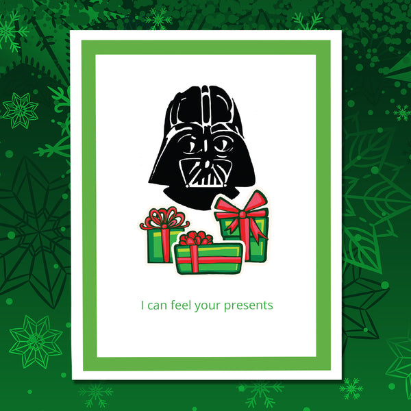 A greeting card featuring a black Darth Vader head above three green gift boxes with red bows, with the text 'I can feel your presents' in green at the bottom. The card has a white center, a green border, and is shown against a green snowflake background.