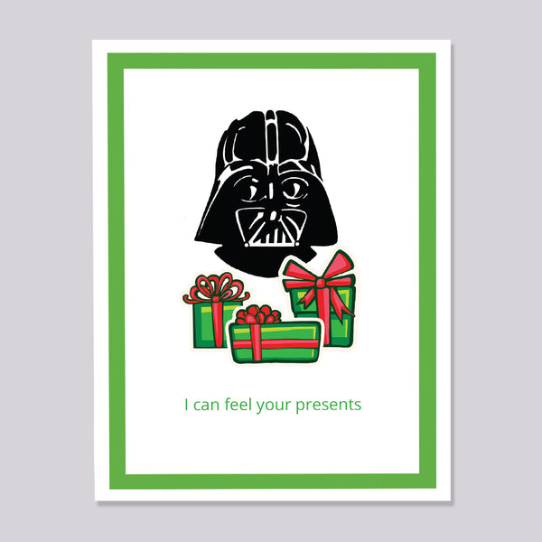 A greeting card featuring a black Darth Vader head above three green gift boxes with red bows, with the text 'I can feel your presents' in green at the bottom. The card has a white center, a green border, and is shown against a light gray background.