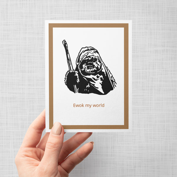 A hand holding a greeting card featuring a black-and-white illustration of a Star Wars Ewok holding a staff, with the text 'Ewok my world' in brown below. The print has a white center and a tan inner border shown against a neutral light gray background.