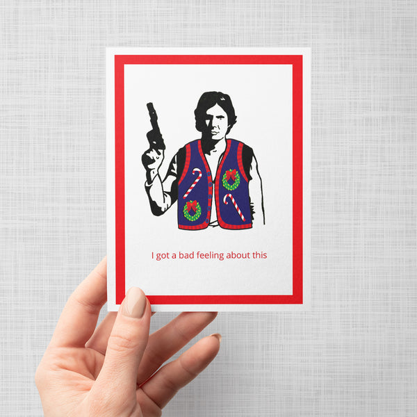 A hand holding a greeting card featuring a stylized black-and-white illustration of Han Solo wearing a Christmas sweater vest with candy canes and wreaths, holding a gun. Below the illustration, red text reads, 'I got a bad feeling about this.