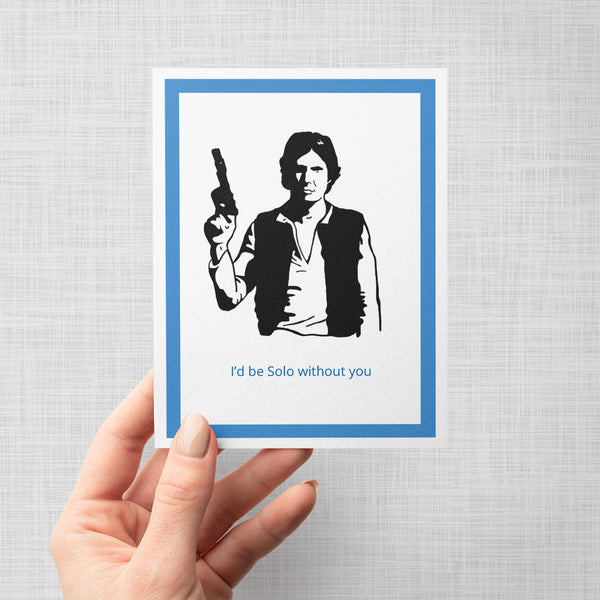 A hand holding a greeting card featuring a black-and-white illustration of Han Solo holding a gun, with the blue text 'I'd be Solo without you' at the bottom. The card is centered on a light gray background with a blue inner border.