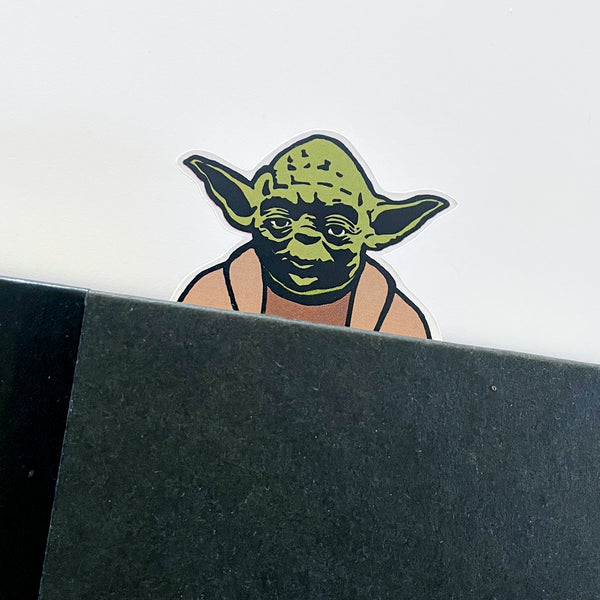 Illustrated die cut yoda character from star wars as a bookmark. His head peeks out of the top of a black book.