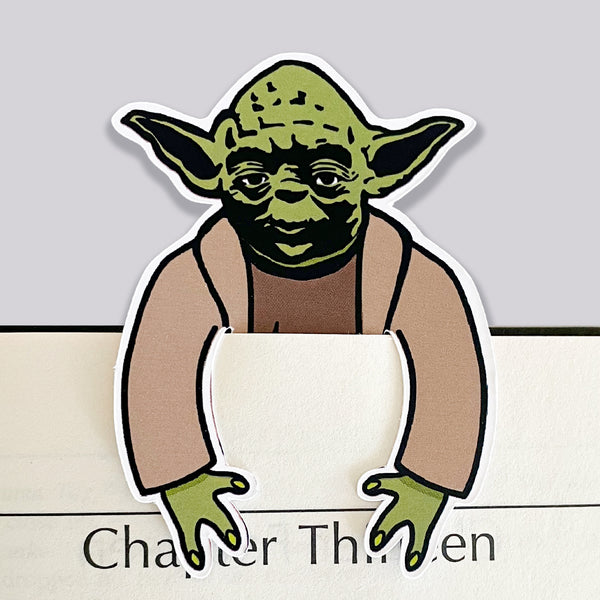 Illustrated die cut yoda character from star wars as a bookmark. His arms hook over the front of the page of a book.