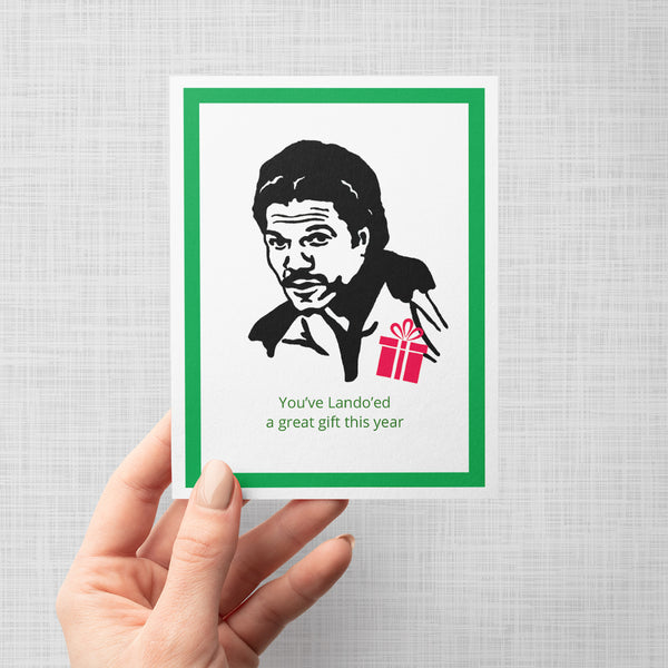 A hand holding a greeting card featuring a black-and-white illustrated portrait of a lando Calrissian, a small red gift box, and the text 'You've Lando'ed a great gift this year' on a white card with a green border, shown against a light gray background.