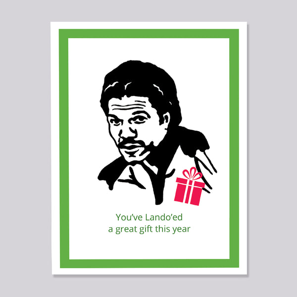 A greeting card featuring a black-and-white illustrated portrait of a lando Calrissian, a small red gift box, and the text 'You've Lando'ed a great gift this year' on a white card with a green border, shown against a light gray background.