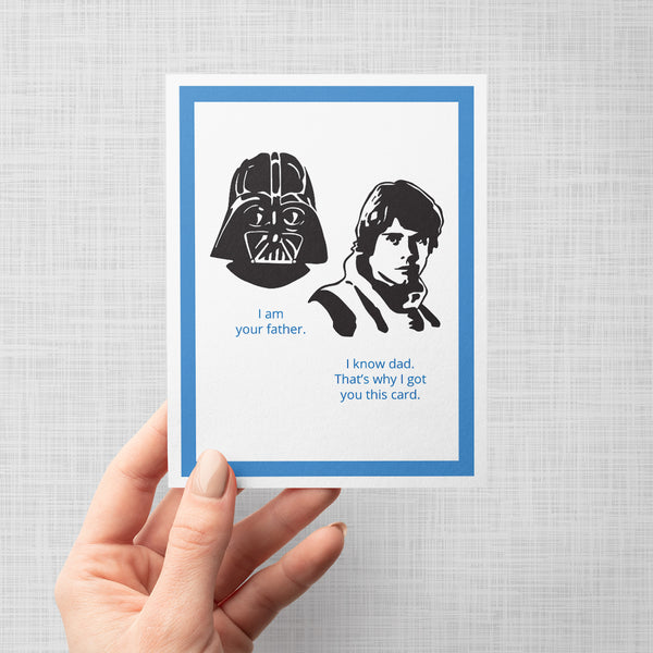 A hand holding a greeting card with a black and white Star Wars-themed illustration showing Darth Vader and Luke Skywalker with the text 'I am your father. I know dad. That's why I got you this card' on a gray background