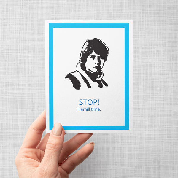A hand holding a greeting card featuring a black-and-white portrait of Luke Skywalker above the text 'STOP! Hamill time.' in blue, framed by a bright blue border on a light gray background.