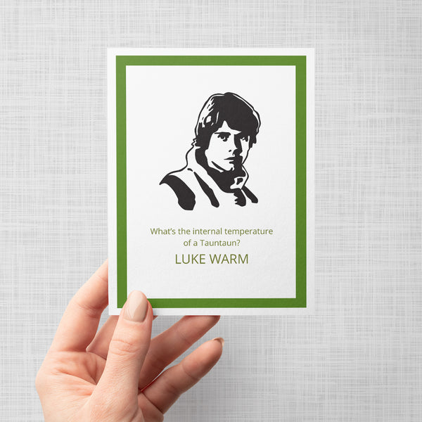 A hand holding a greeting card featuring a black and white graphic portrait of Luke Skywalker with green text reading 'What's the internal temperature of a Tauntaun? LUKE WARM'. The card has a white border and is framed by a dark green inner border and a light gray outer background.