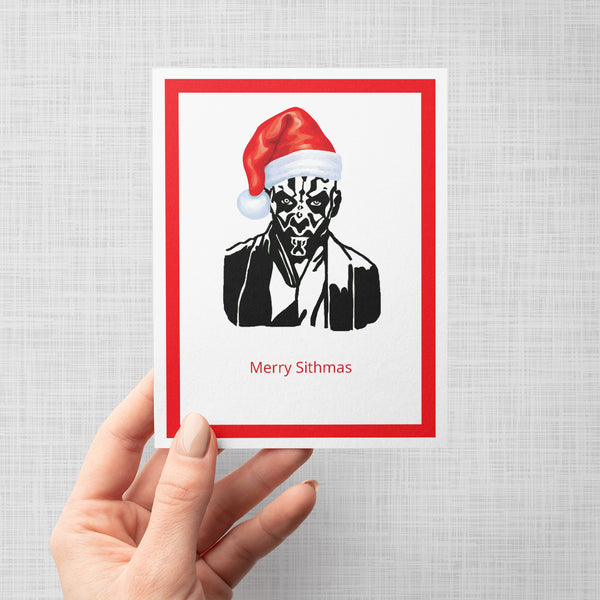 A hand holding a greeting card featuring a stylized black-and-white portrait of Darth Maul wearing a red Santa hat, with the text 'Merry Sithmas' below. The card has a white border with a thick red inner border and is shown against a light gray background.