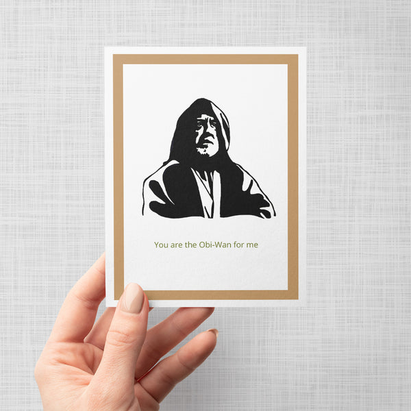 A hand holding a greeting card featuring a black-and-white stylized portrait of Obi-Wan Kenobi with the text 'You are the Obi-Wan for me' below, framed by a tan border on a light gray background.