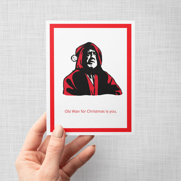 A hand holding a greeting card featuring a stylized graphic portrait of Obi Wan wearing a red Santa hat and robe on a white background, framed by a bold red border. Below the portrait, the text reads 'Obi Wan for Christmas is you.'