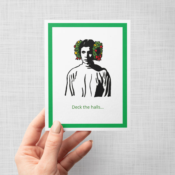 A hand holding a greeting car featuring a stylized black-and-white portrait of Princess Leia with green holiday wreaths on the sides of her head, with the text 'Deck the halls. 