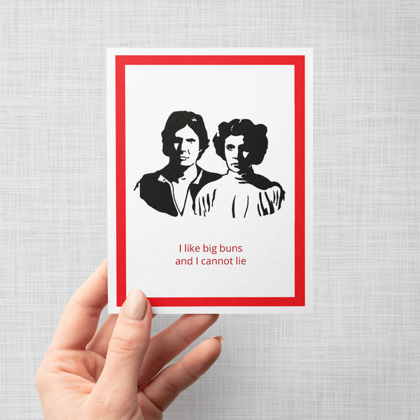 A hand holding a greeting card featuring a black-and-white illustration of Princess Leia and Han Solo, with red text below reading 'I like big buns and I cannot lie.' The print has a white border and a wide dark red frame, shown against a light gray background.