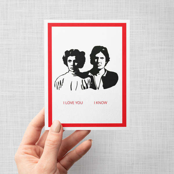 A hand holding a greeting card featuring a black-and-white illustration of Princess Leia and Han Solo, with red border and the text 'I LOVE YOU I KNOW' in red at the bottom, shown against a light gray background.