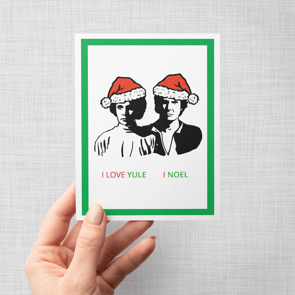 A hand holding a greeting card featuring a minimalist black-and-white illustration of Princess Leia and Han Solo wearing red Santa hats, with the text 'I LOVE YULE | I NOEL' at the bottom. The card has a white border and a green inner border, shown against a light gray background.