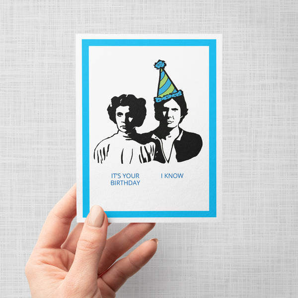 A hand holding a greeting card with black and white illustrations of Princess Leia and Han Solo wearing party hats, text 'It's your birthday' and 'I know', on a gray background.