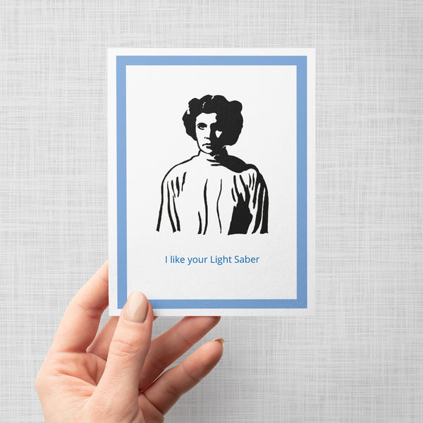 A hand holding a greeting card featuring a black illustration of a Princess Leia with a light saber, bordered by a blue frame on a white background, with the text 'I like your Light Saber' printed in blue at the bottom.