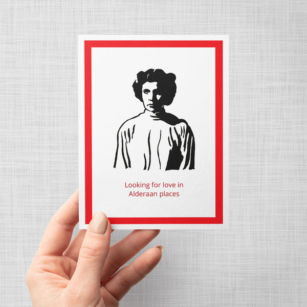 A hand holding a greeting card featuring a black and white illustration of Princess Leia, bordered by a red frame, with the red text 'Looking for love in Alderaan places' at the bottom, shown against a neutral gray background.