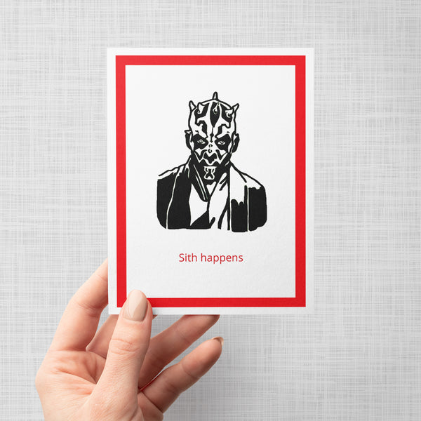A hand holding a greeting card featuring a black ink illustration of Darth Maul with the text 'Sith happens' in red below. The card is framed by a red inner border, set against a neutral light gray background.