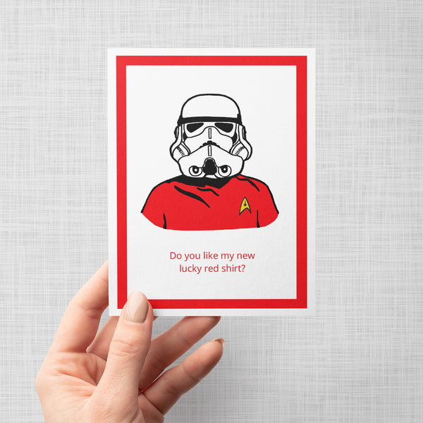 A hand holding a greeting card featuring a stylized illustration of a Star Wars stormtrooper wearing a red Star Trek uniform shirt with a yellow delta insignia. Below the illustration, the text reads 'Do you like my new lucky red shirt?'.