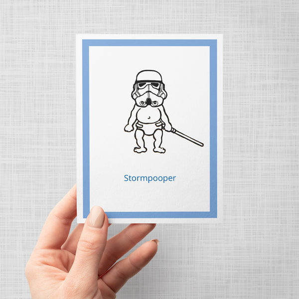 A hand holding a greeting card featuring a black line drawing of a baby dressed as a Star Wars stormtrooper holding a lightsaber, with the word 'Stormpooper' in blue text below. The card has a white center, a blue border, and is shown against a light gray background.