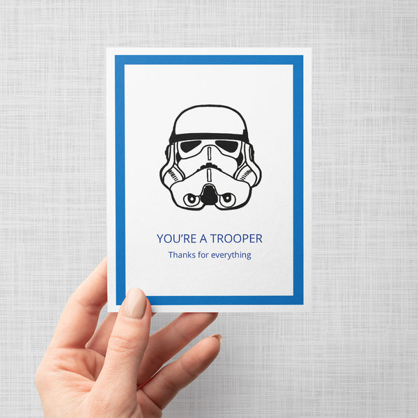 A hand holding a greeting card featuring a black line art illustration of a Star Wars stormtrooper helmet on a white background, framed by a blue border. Below the illustration, the text reads 'YOU'RE A TROOPER' and 'Thanks for everything' in blue.