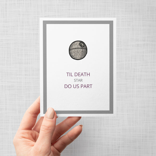 A hand holding a greeting card featuring a black and gray line drawing of the Star Wars Death Star above the text 'TIL DEATH STAR DO US PART' in purple lettering, set against a white background with a gray border.