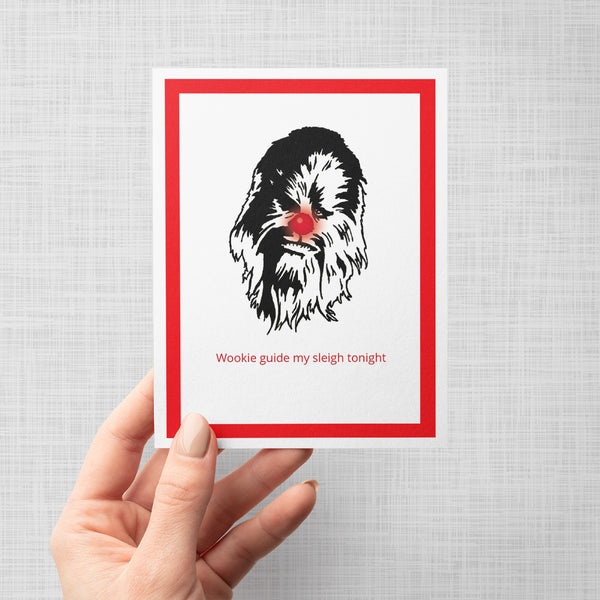 A hand holding a greeting card featuring a black line-art illustration of Chewbacca's face with the red text 'Wookie guide my sleigh tonight' underneath, framed by a bold red border on a white background, shown against a light gray backdrop.