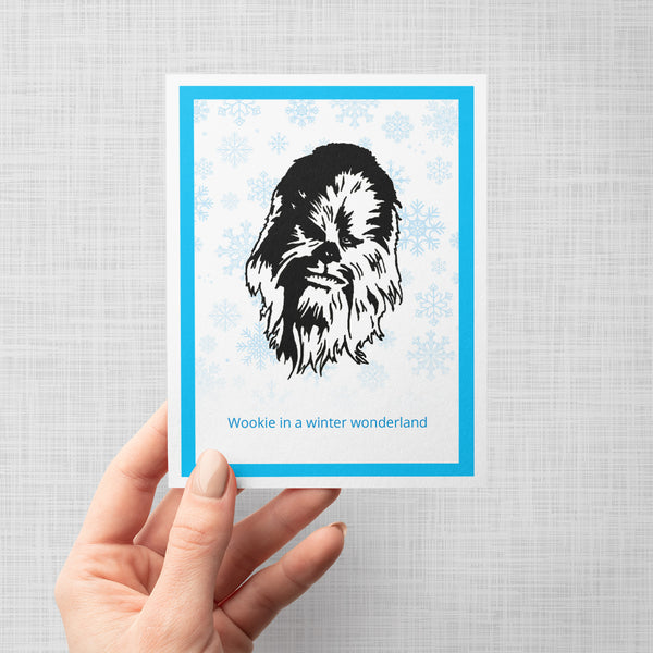 A hand holding a greeting card featuring a black-and-white illustration of Chewbacca centered on a white background with light blue snowflakes, framed by a bright blue border. The text 'Wookie in a winter wonderland' appears in blue at the bottom.