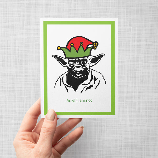 A hand holding a greeting card featuring a black-and-white illustration of Yoda wearing a red and green elf hat, with the text 'An elf I am not' below. The print has a white center, a green border, and is shown against a light gray background.