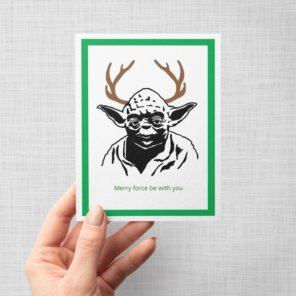 A hand holding a greeting card featuring a stylized black-and-white portrait of Yoda with brown antlers and green text reading 'Merry force be with you' at the bottom, framed by a green border on a white print background against a light gray backdrop.