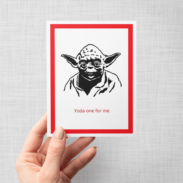 A hand holding a greeting card featuring a black line art illustration of Yoda with the text 'Yoda one for me' in red below. The print has a white border, a thick magenta-pink inner border, and is displayed against a neutral light gray background.