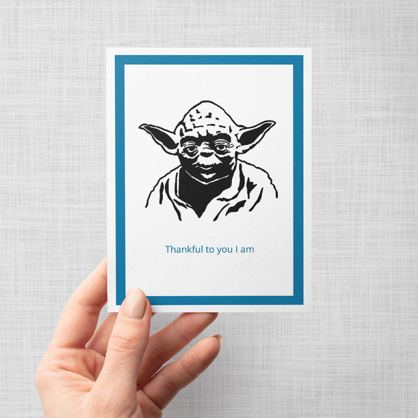 A hand holding a greeting card featuring a black ink illustration of Yoda with the text 'Thankful to you I am' in blue below it, framed by a white border and a teal inner border on a light gray background.