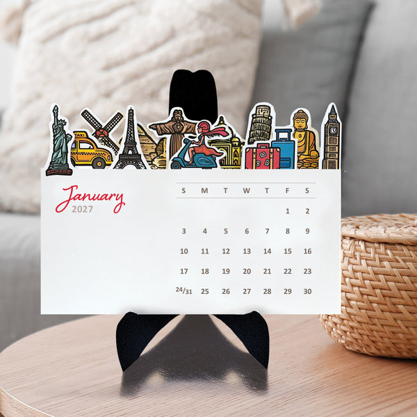 A 2027 desk calendar page featuring colorful travel-themed illustrations such as the statue of liberty, the taj mahal, pyramds, big ben and the statue of liberty along the top, displayed on a black stand on a wooden table with a woven basket and soft-focus indoor background.