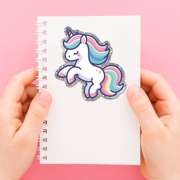 Glittery unicorn die cut vinyl sticker with a colorful mane on a notebook held by hands against a pink background